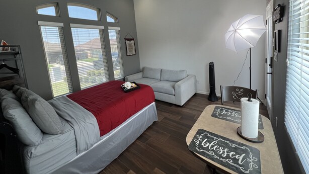 A bedroom with a bed, a gray sofa, a red blanket, a standing fan, and a window with blinds.