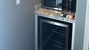 A kitchenette with a microwave, coffee maker, and Keurig coffee maker on the counter. There is a framed picture on the wall and a shelf with various items.