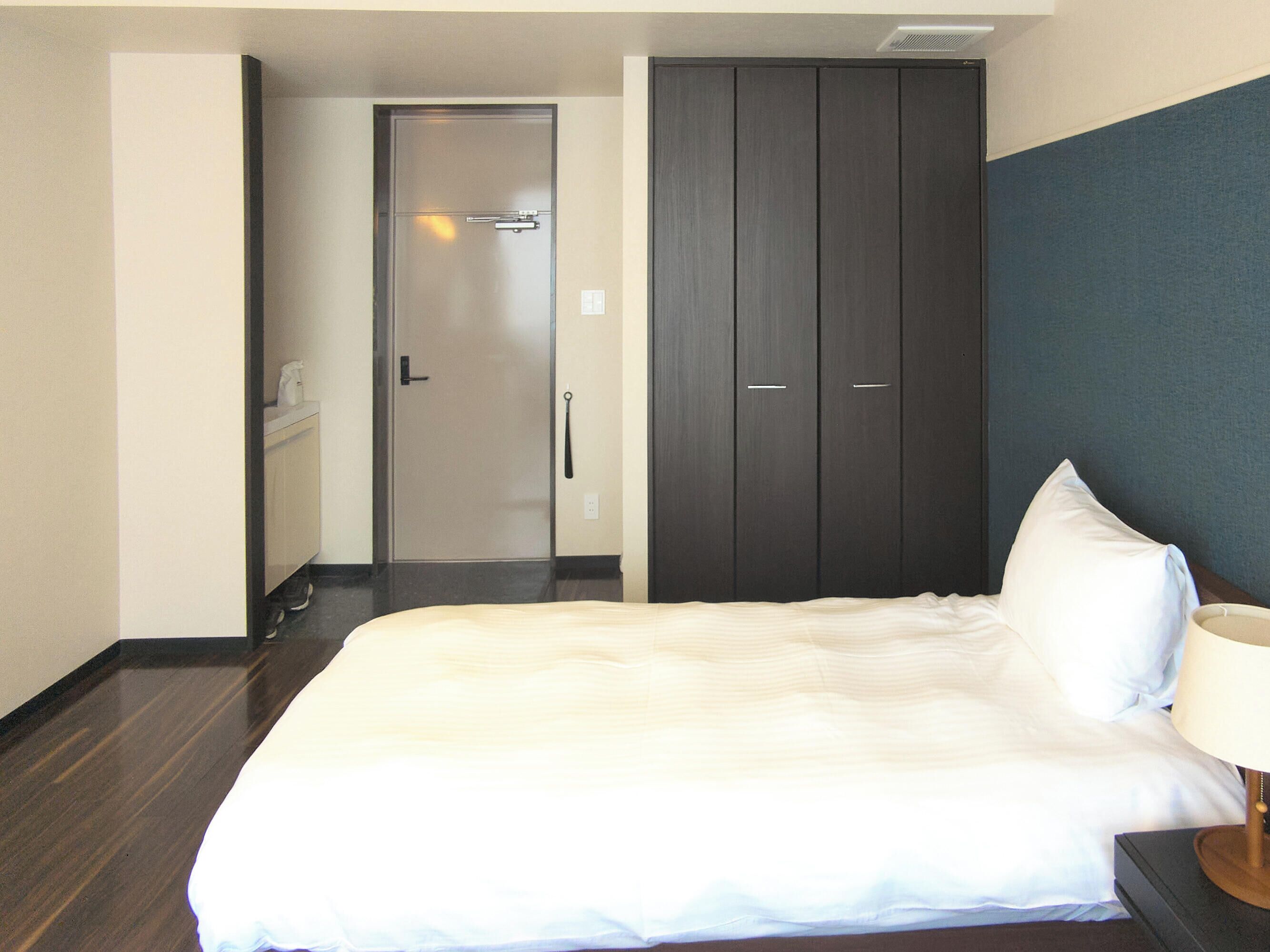 Habitación de hotel con cama, mesita de noche, lámpara, armario y puerta que da a un baño.