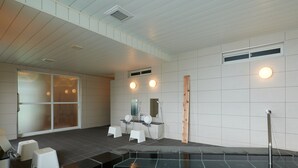 Zona d'una piscina interior moderna amb una vista clara, dos tamborets blancs, un piló de fusta i una zona de dutxes amb un mirall i dues làmpades rodones.