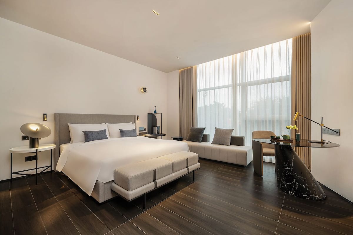 Quarto de hotel moderno com uma cama grande, um sofá, uma mesa pequena e vista da cidade através de amplas janelas.