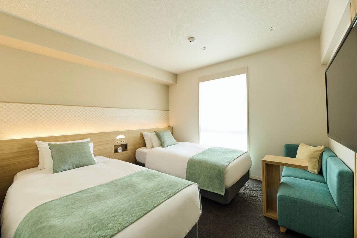 A hotel room with two beds, a small sofa, a wooden nightstand, and a large window providing natural light.