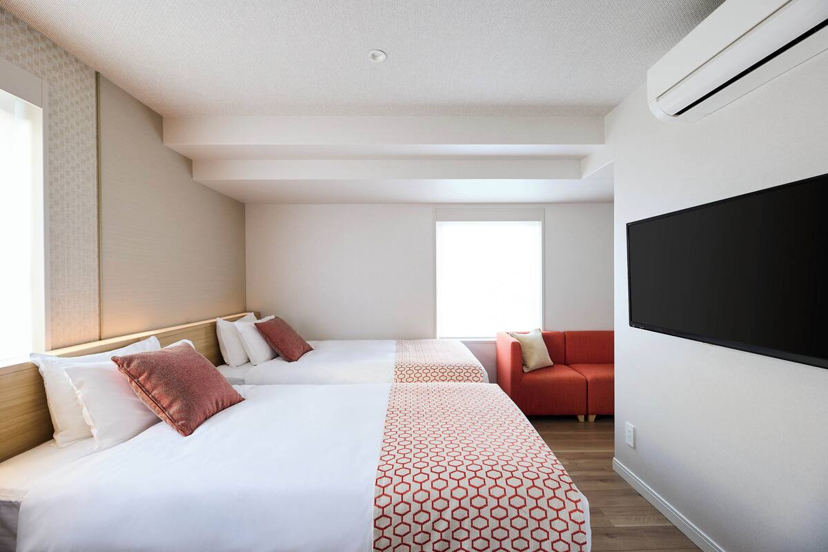 A hotel room with two beds, a red sofa, a large TV, and a window providing natural light.