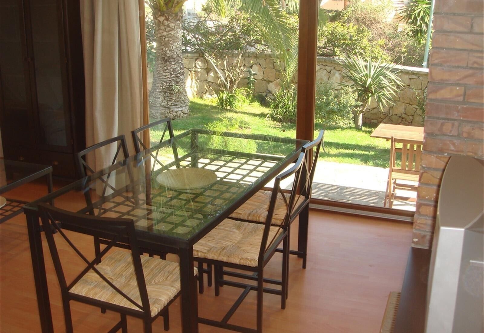 Une table de salle à manger avec un plateau en verre, quatre chaises, un mur en pierre et une vue sur un jardin.