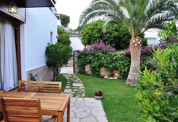 A patio with wooden furniture, a stone pathway, and lush greenery including a palm tree and flowering plants. - Holiday Villa in Tarragona with Open Terrace, Garden & Wi-Fi (Tarragona)