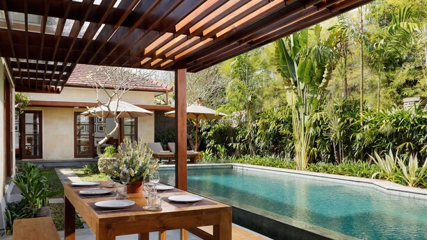 A wooden dining table set with plates, glasses, and a vase, overlooking a swimming pool with lush greenery. - Villa Perere9 by Nakula (Canggu)