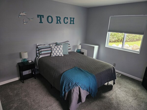 Une chambre à coucher comprenant un lit, une table de chevet, une lampe, une fenêtre avec des stores et un mur sur lequel est écrit le mot « TORCH » au-dessus du lit.
