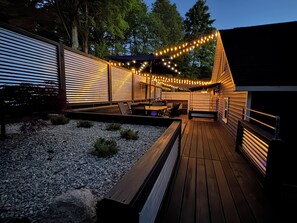 A wooden deck with string lights, seating, and a small garden area. - Spacious home near Torch Lake w/custom back patio, steps away from public access (Alden)