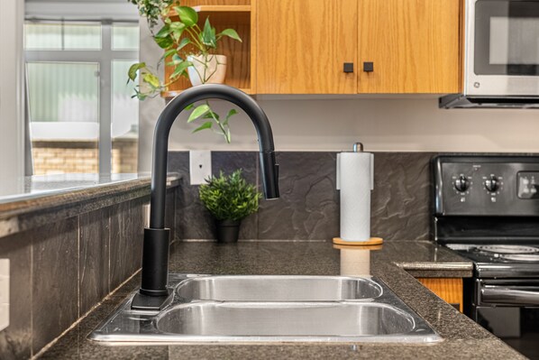 A kitchen with a stainless steel sink, a black faucet, a microwave above the stove, and wooden cabinets. - River Downtown Condo, A/C, Underground Parking, Saddledome & Stampede (Calgary)
