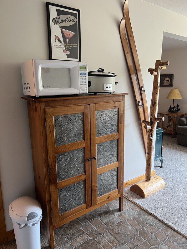 A wooden cabinet with a microwave and a slow cooker on top, a framed poster on the wall, and a wooden ladder leaning against the wall.