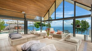 A modern, open-plan living area with a large glass wall offering a sea view, a pool, and outdoor seating. - Villa Saengootsa (Kamala)
