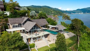 A modern house with a large swimming pool, surrounded by lush greenery and overlooking a body of water. - Villa Saengootsa (Kamala)