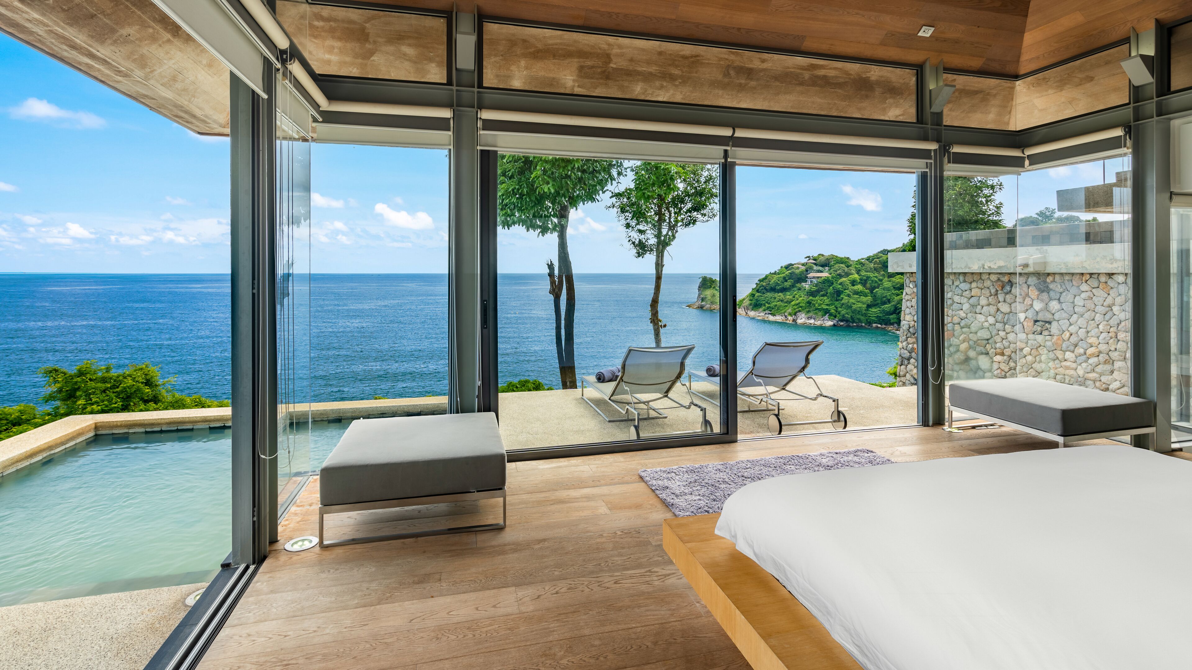 A modern bedroom with a large bed, a glass door leading to a balcony with a view of the sea and a stone wall.