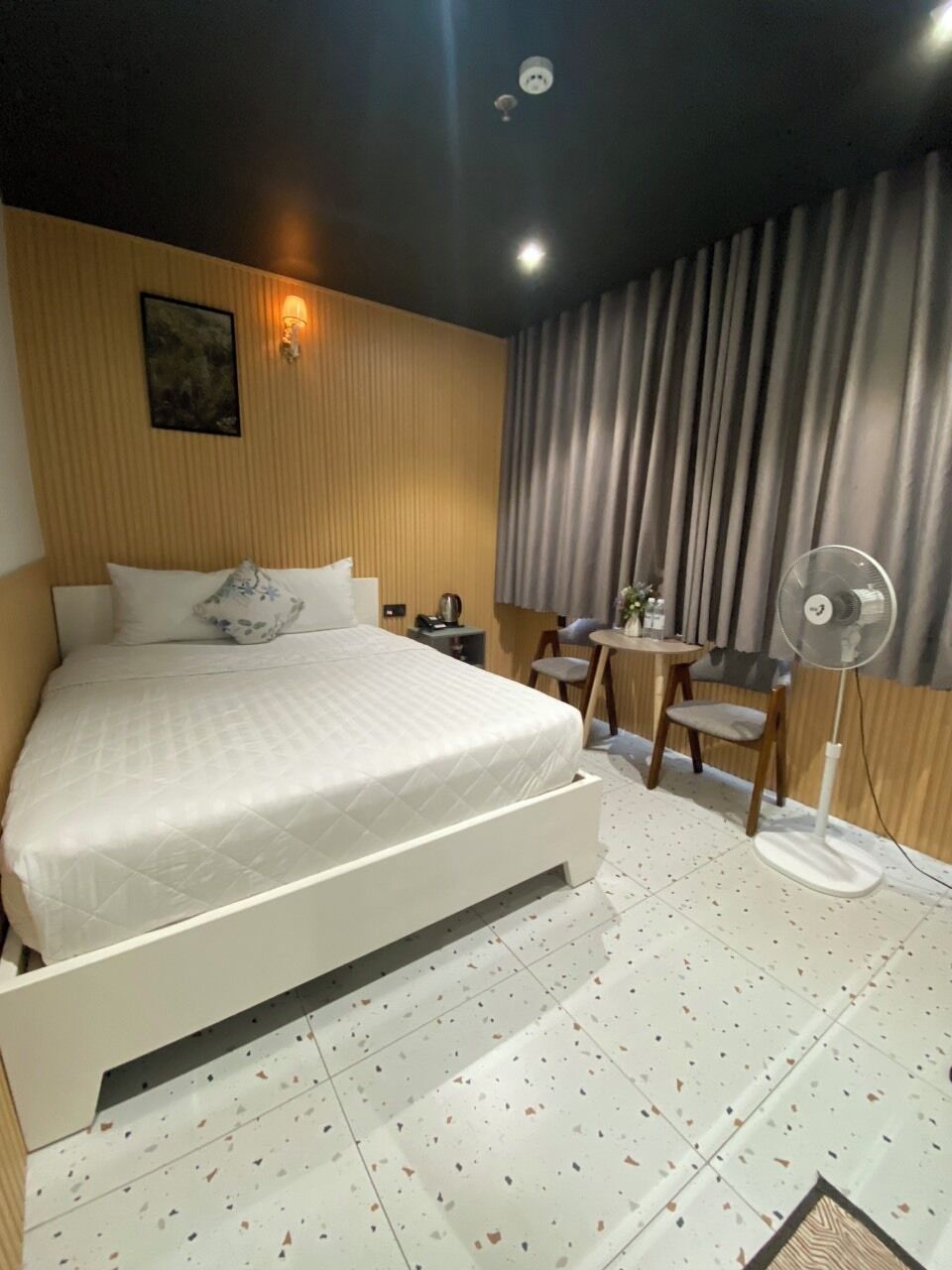 Habitación de hotel con cama, escritorio, silla, mesa, ventilador y ventana con cortinas.