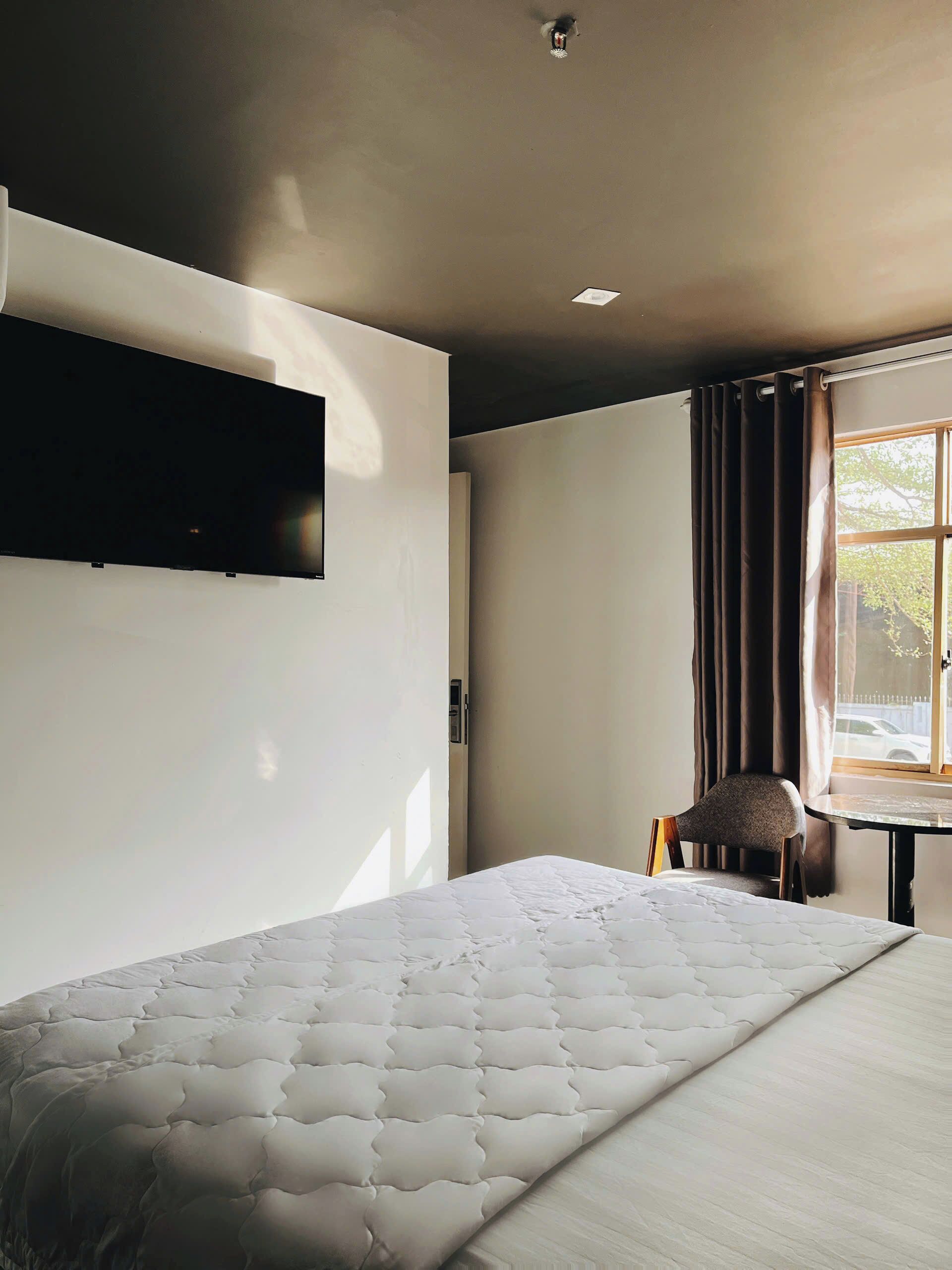 A bedroom with a bed, a chair, a table, a window with curtains, and a wall-mounted television.