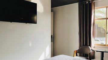 A bedroom with a bed, a chair, a table, a window with curtains, and a wall-mounted television.