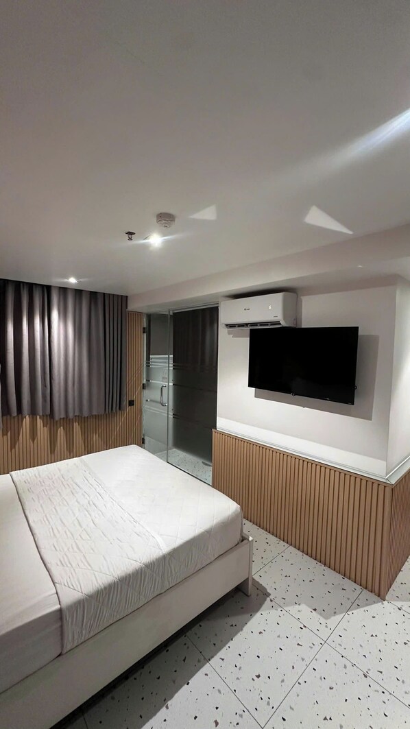A modern hotel room with a bed, a flat-screen TV mounted on the wall, and a sliding glass door leading to a bathroom.