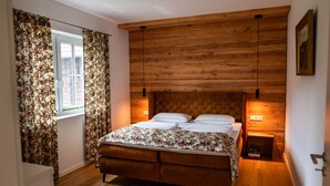 Une chambre à coucher avec une tête de lit en bois, un lit recouvert d’une literie à motifs floraux, une fenêtre ornée de rideaux à fleurs et une table de chevet avec une lampe.
