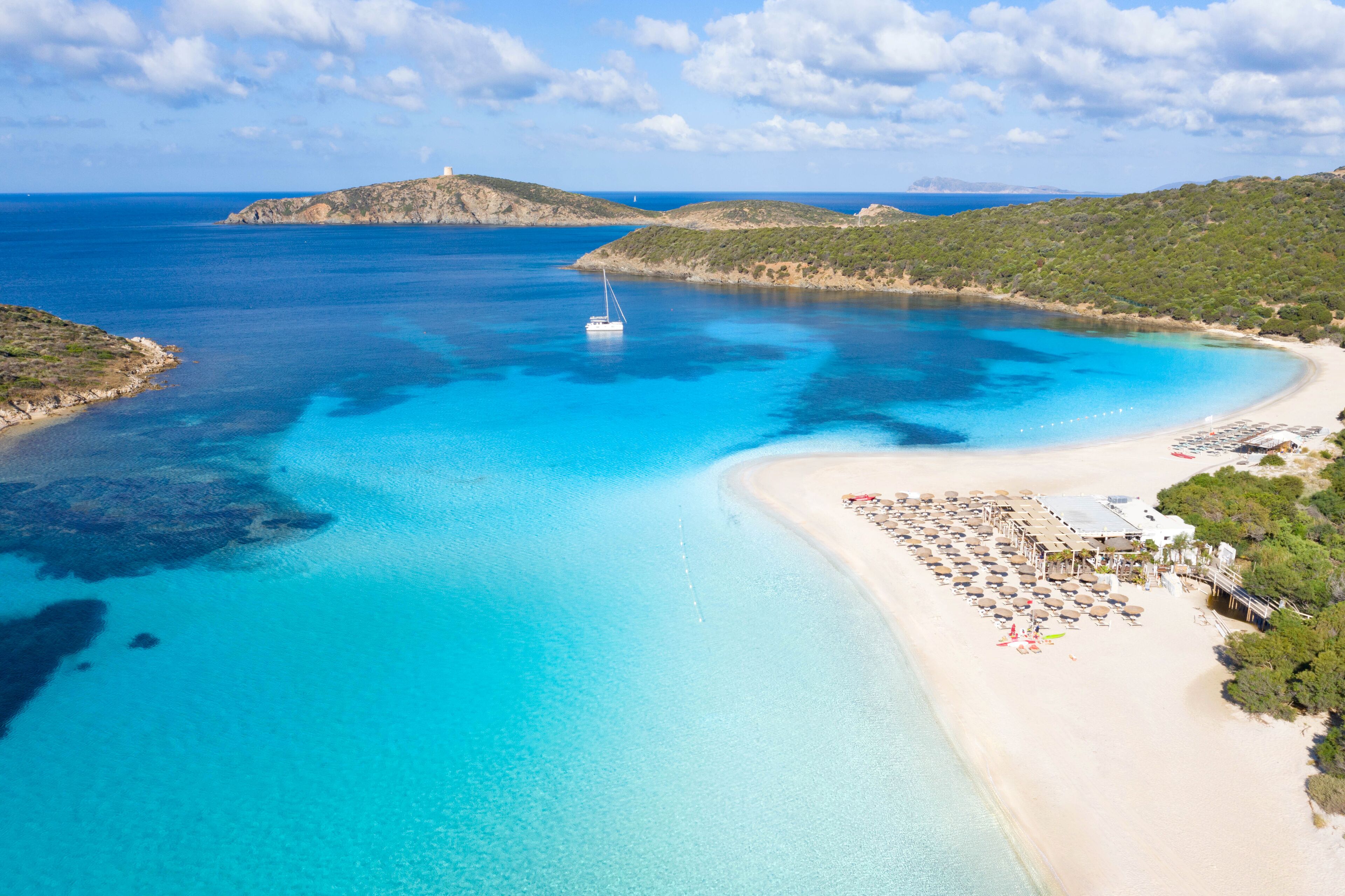 Una playa costera con un bote, aguas azules cristalinas y un área de resort con sombrillas y sillones reclinables.