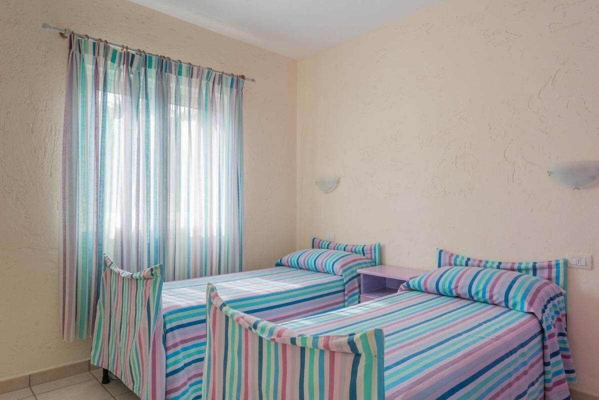 Una habitación con dos camas, una ventana con cortinas, una mesita y una lámpara fijada a la pared.
