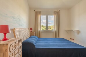 Un dormitorio con cama, cómoda de madera, lámpara roja, ventana con cortinas y vista a la vegetación.