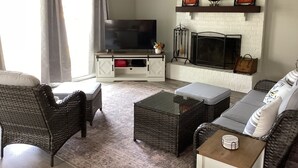Un salon avec un téléviseur, une cheminée, des meubles en osier et un mur en briques blanches.