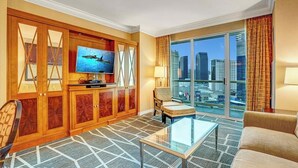 Ein Hotelzimmer mit einem großen Fernseher, einem Glastisch, einer Couch, einem Sessel und Blick auf die Stadt durch große Fenster.