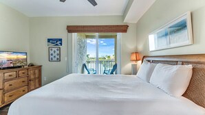 A hotel room with a large bed, a TV on a wooden dresser, a balcony with chairs, and a painting on the wall. - Summerhouse 101 - Pearl of the Gulf (Port St. Joe)