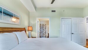 A bedroom with a large bed, a nightstand with a lamp, a door leading to a bathroom, and a ceiling vent. - Summerhouse 101 - Pearl of the Gulf (Port St. Joe)