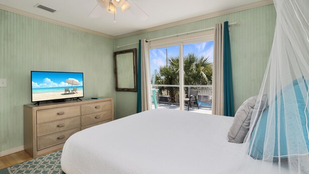 Een slaapkamer met een bed, een televisie op een dressoir, een raam met gordijnen en uitzicht op palmbomen.