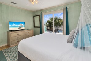 Quarto com cama, televisão em uma cômoda, janela com cortinas e vista para palmeiras lá fora.