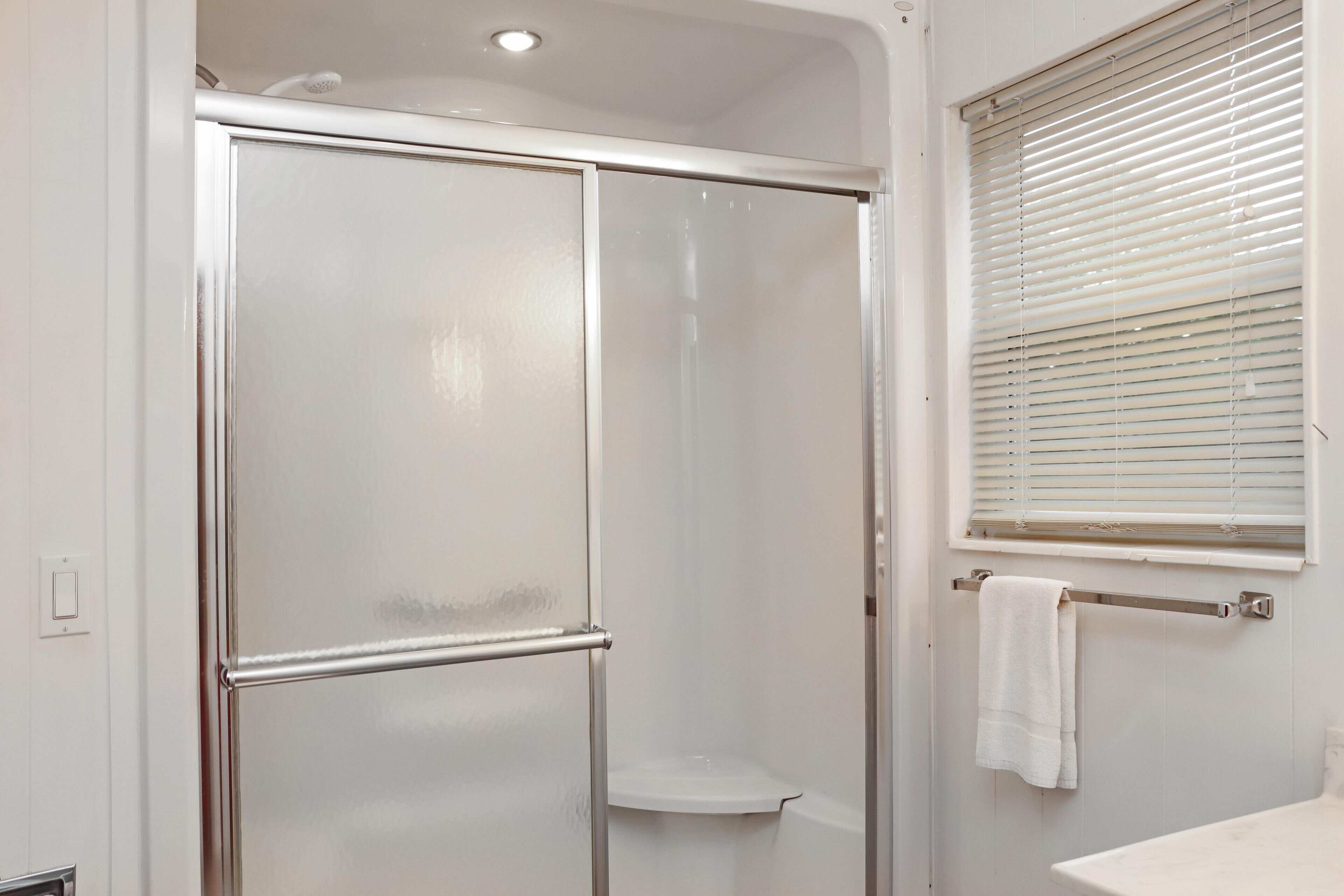 Een badkamer met een douchecabine van glas, een wit wastafeltje en een raam met jaloezieën.