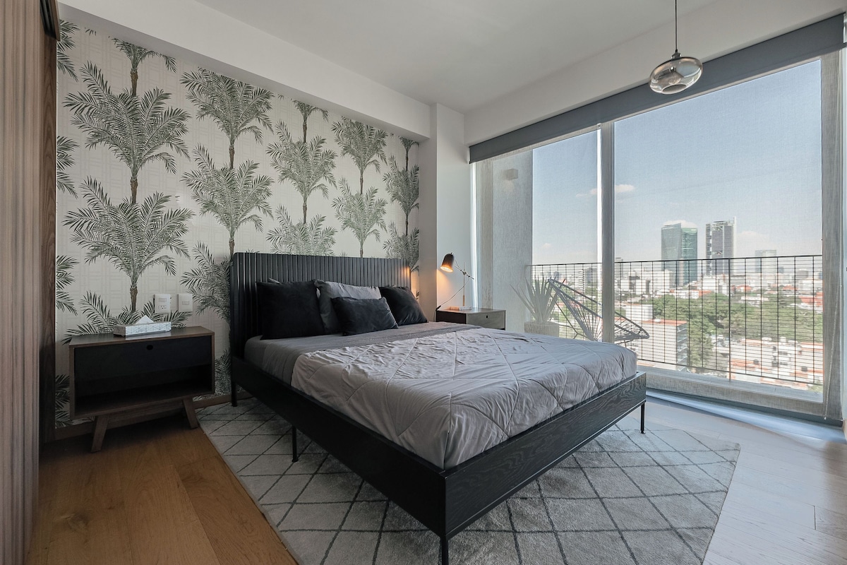 Un dormitorio moderno con una cama grande, una mesita de noche, un televisor y vistas a la ciudad a través de amplios ventanales.