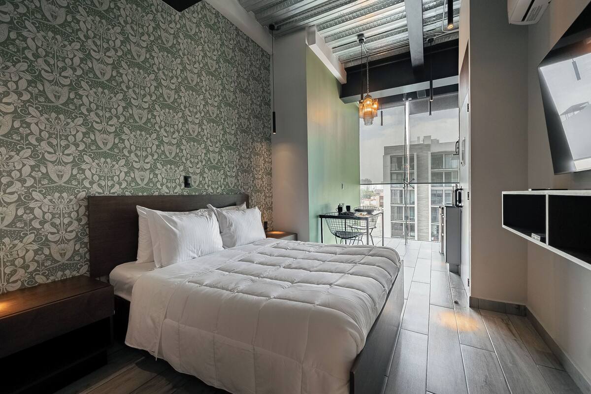A modern bedroom with a large bed, a patterned wallpaper, a small table with chairs, and a view through a glass door.