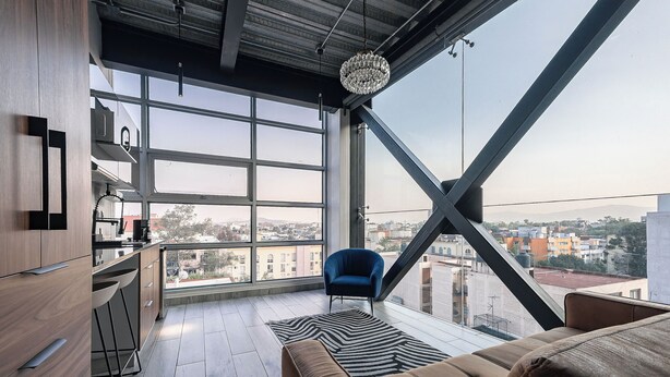 A modern living room with a large window, a blue chair, a sofa, and a kitchen area with wooden cabinets and a bar stool. - Mr. Herold homes (Mexico City)
