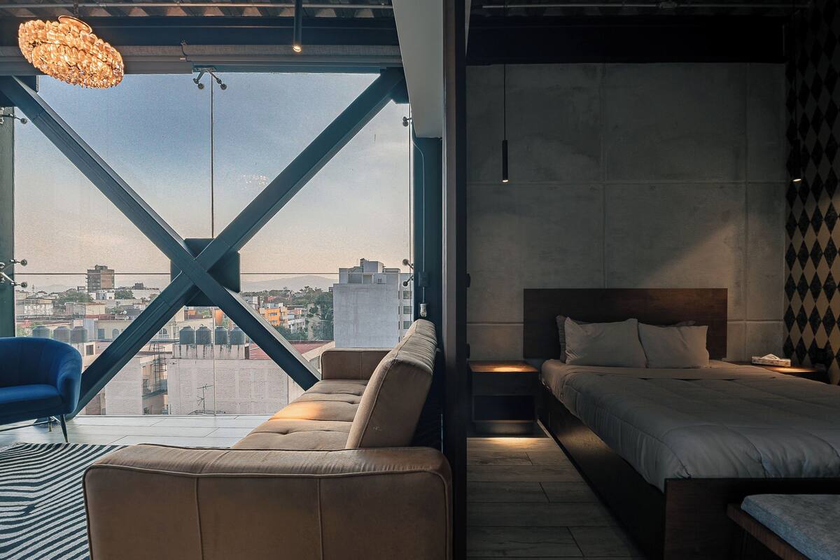 A modern bedroom with a large window, a sofa, a bed, and a blue chair.