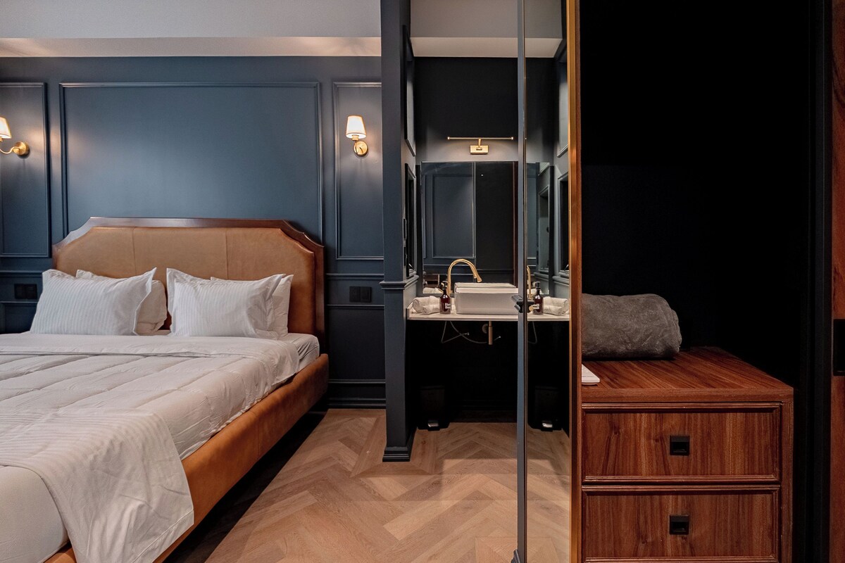 A modern hotel room with a large bed, a wooden nightstand, a mirror, and a built-in vanity with a sink.