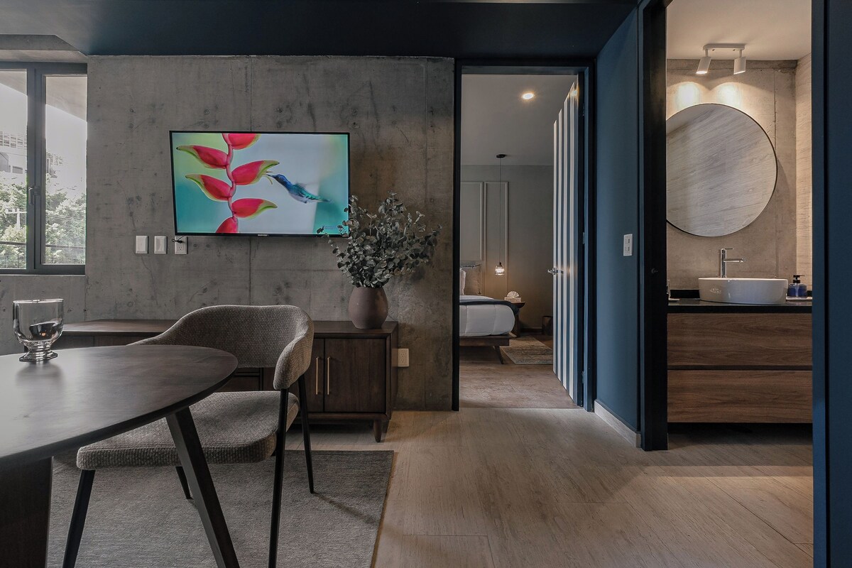 Moderni hotellihuone, jossa on ruokailutila, suuri litteänäyttöinen televisio, ruukkukasvi ja pyöreä peili pesualtaan yläpuolella.