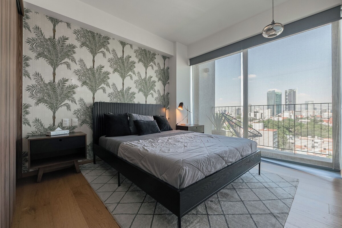 A modern bedroom with a large bed, a nightstand, a TV, and a city view through large windows.