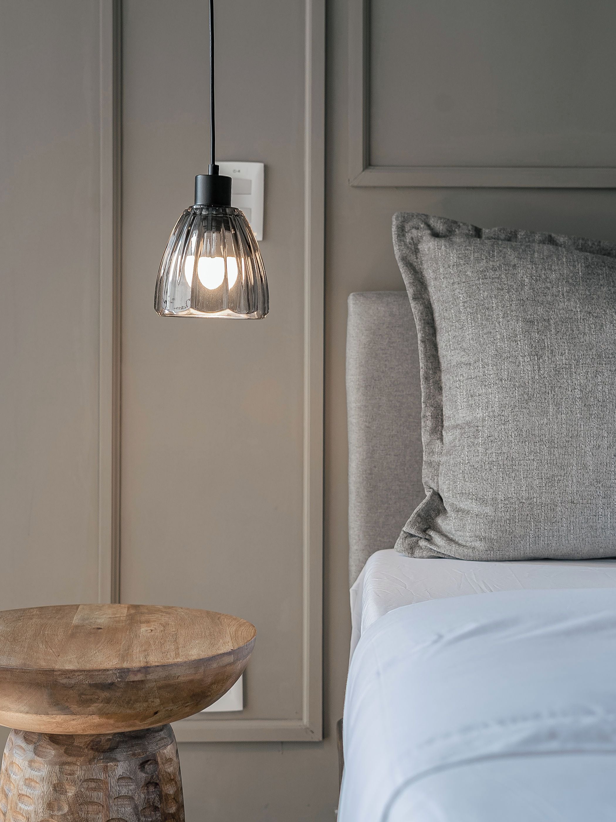 A bedroom with a wooden table, a hanging pendant light, and a bed with a grey pillow.