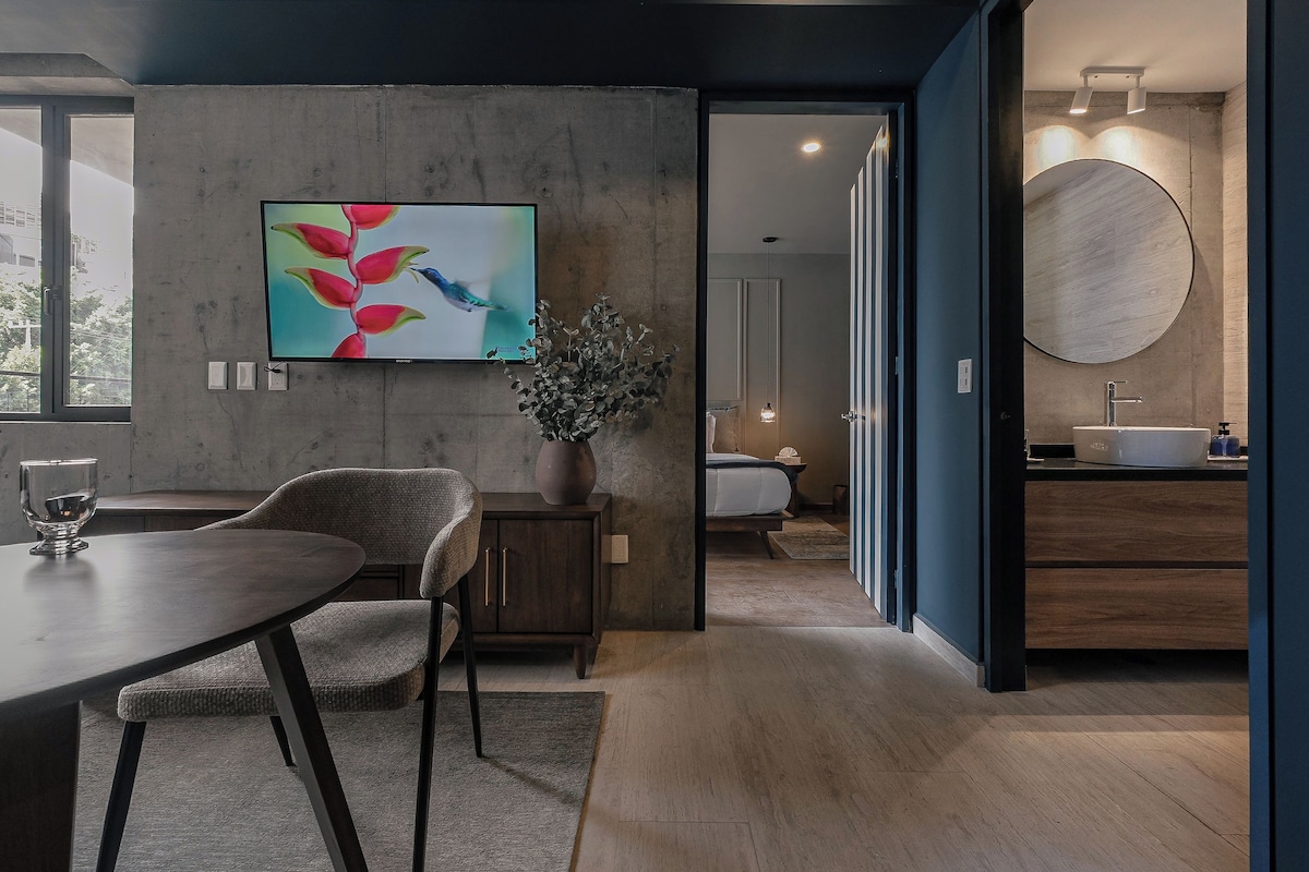 A modern hotel room with a dining area, a large flat-screen TV, a potted plant, and a round mirror above the sink.