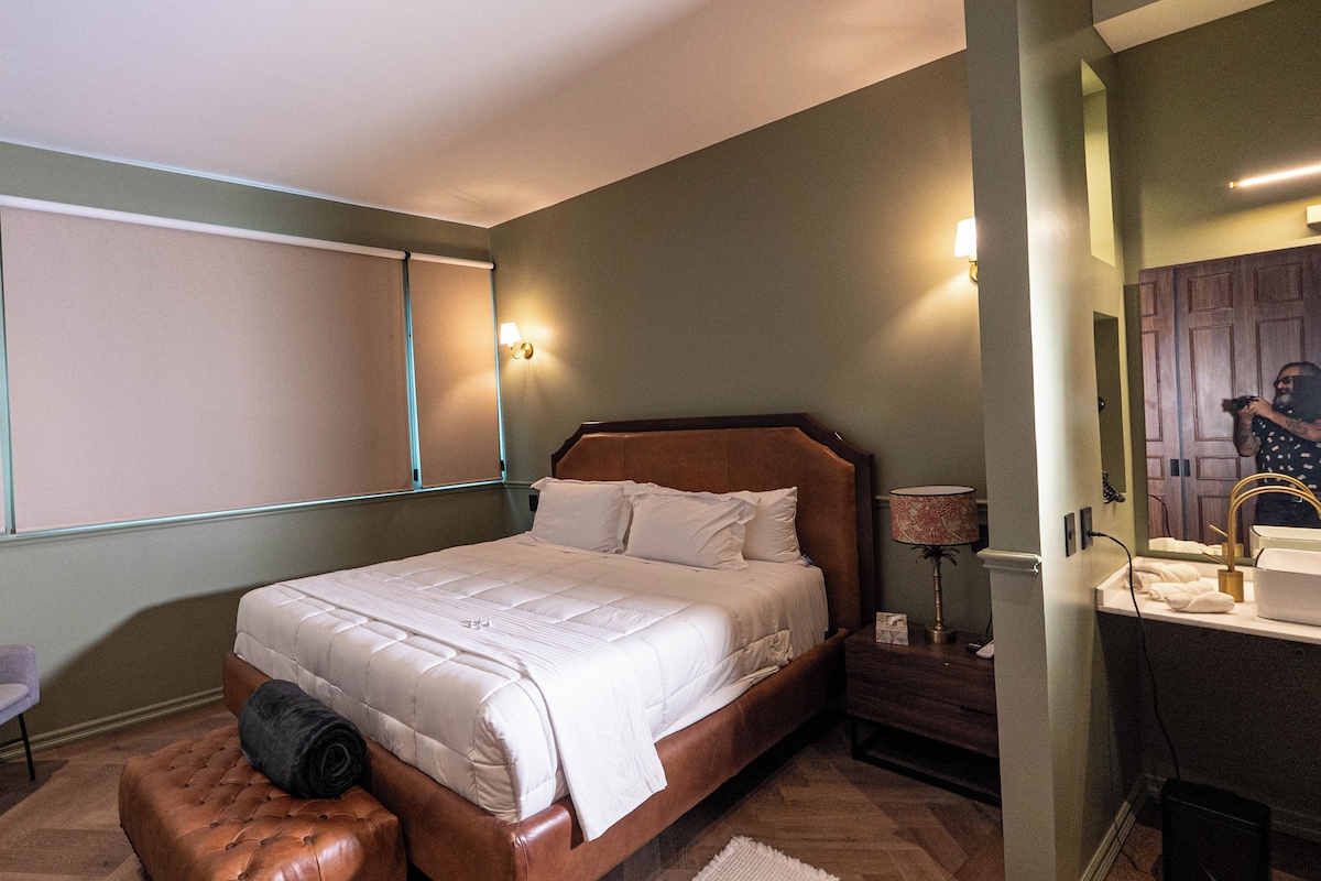 A hotel room with a bed, a chair, a nightstand, a lamp, and a mirror.