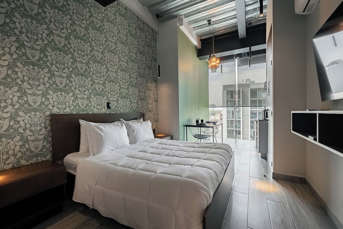 A modern bedroom with a large bed, a patterned wallpaper, a small table with chairs, and a view through a glass door.