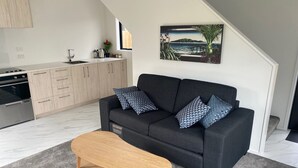 A modern living room with a dark grey sofa, two patterned cushions, and a wooden coffee table. A kitchen with light wood cabinets and a stainless steel stove is visible in the background. - New House in New Brighton, Right by the Beach and Pier! (Christchurch)