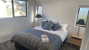 A neatly made bed with a patterned bedspread, two bedside lamps, and a view of a residential area through the window. - New House in New Brighton, Right by the Beach and Pier! (Christchurch)