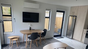 A modern living room with a sofa, a coffee table, a dining table with chairs, a TV mounted on the wall, and a kitchen area with a refrigerator. - New House in New Brighton, Right by the Beach and Pier! (Christchurch)