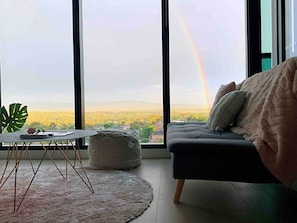A room with a large window, a sofa, a small table, and a plant. - SkyGarden Luxury Apt | Mountain & Sunrise Views (Glen Waverley)