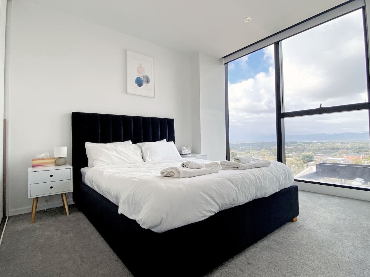 A modern bedroom with a large bed, a nightstand, and a view of the city through a large window.