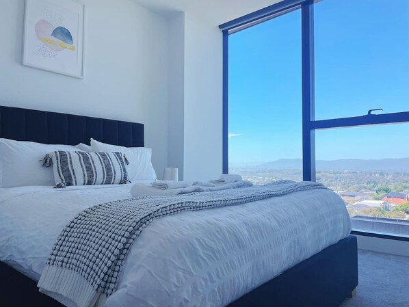 Un dormitorio con un ventanal grande, una cama con ropa de cama blanca y vistas a un paisaje urbano.