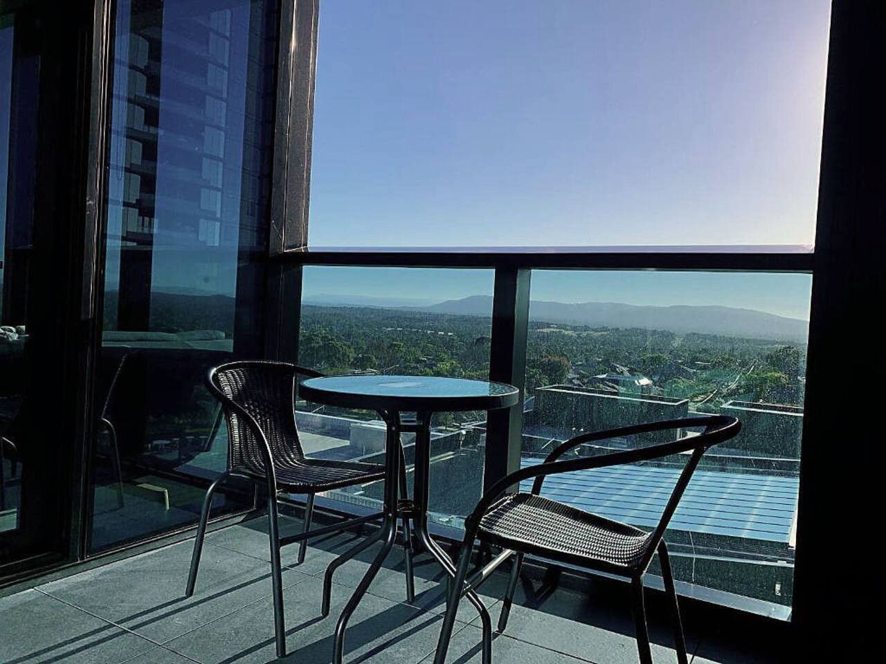 A balcony with two chairs and a table, overlooking a cityscape with buildings and a clear sky.